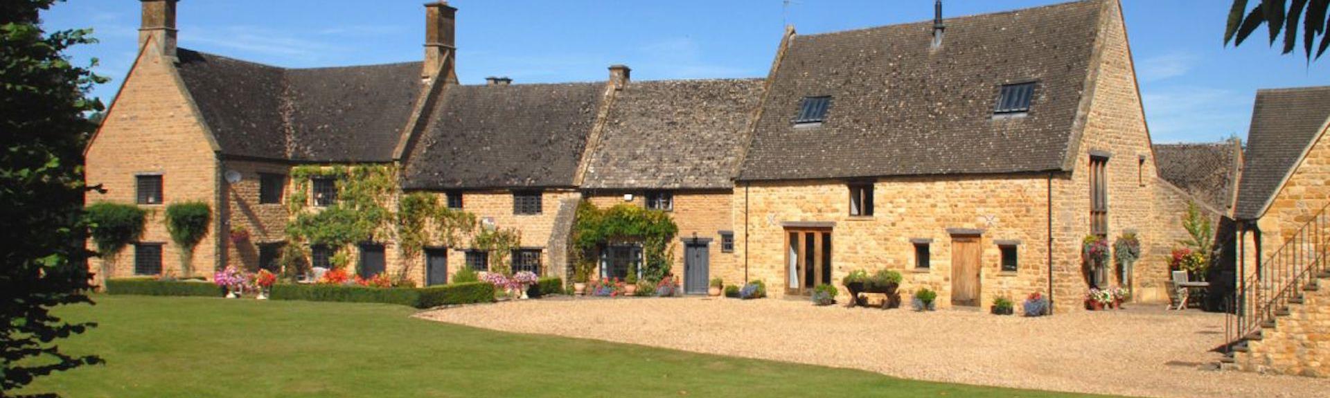 An old Manor House in the Cotswolds with honey-colours bricks and high slate roof overlooks a shingled drive and large lawn.s