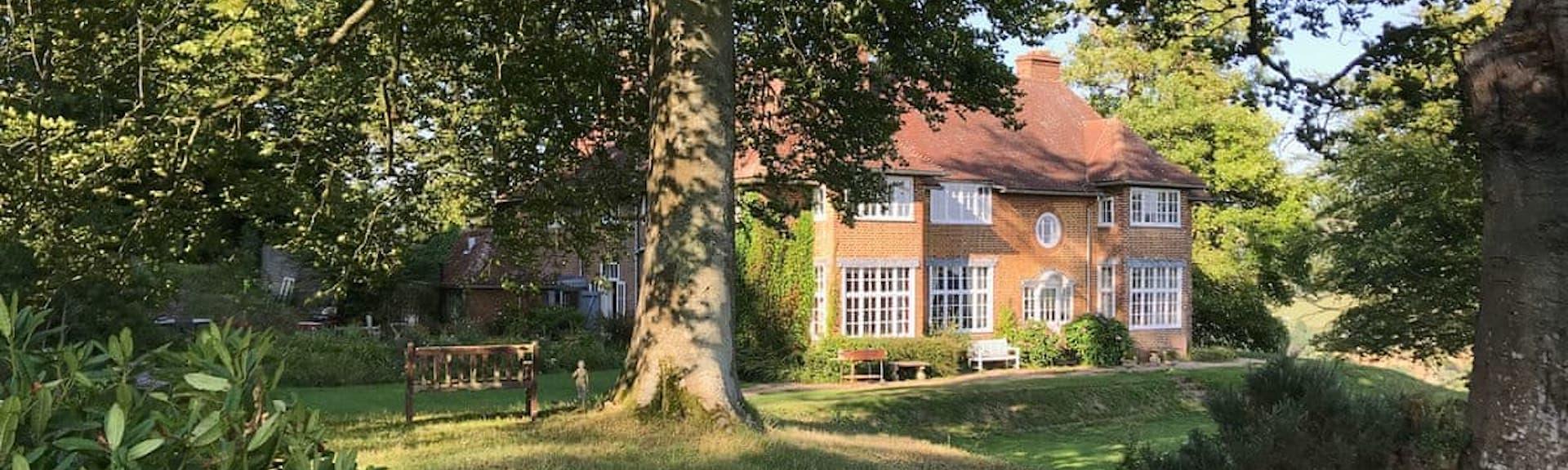 a large detached brick built country house stands in the sunlight within a small woodland glade.