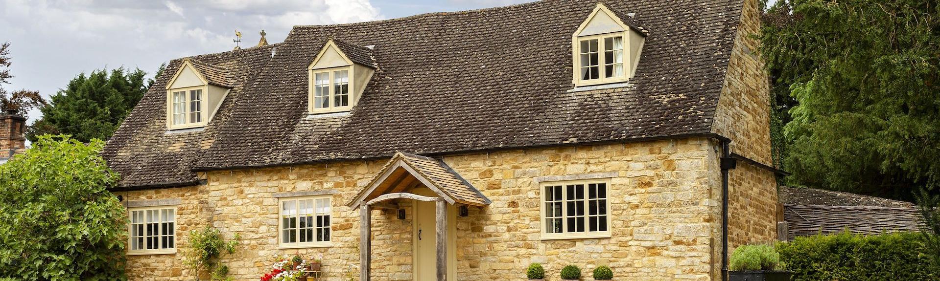 A traditional honeystone cottage in the Cotswolds stands behind a large lawn.