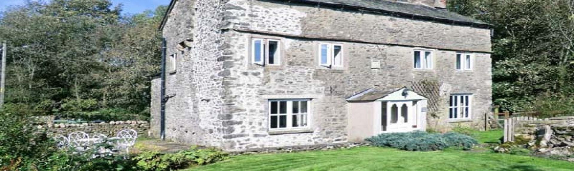 A large double-fronted stone holiday cottage in Kirkby Lonsdale overlooks a newly-mown lawn. Trees and large bushes are to the rear of the house.