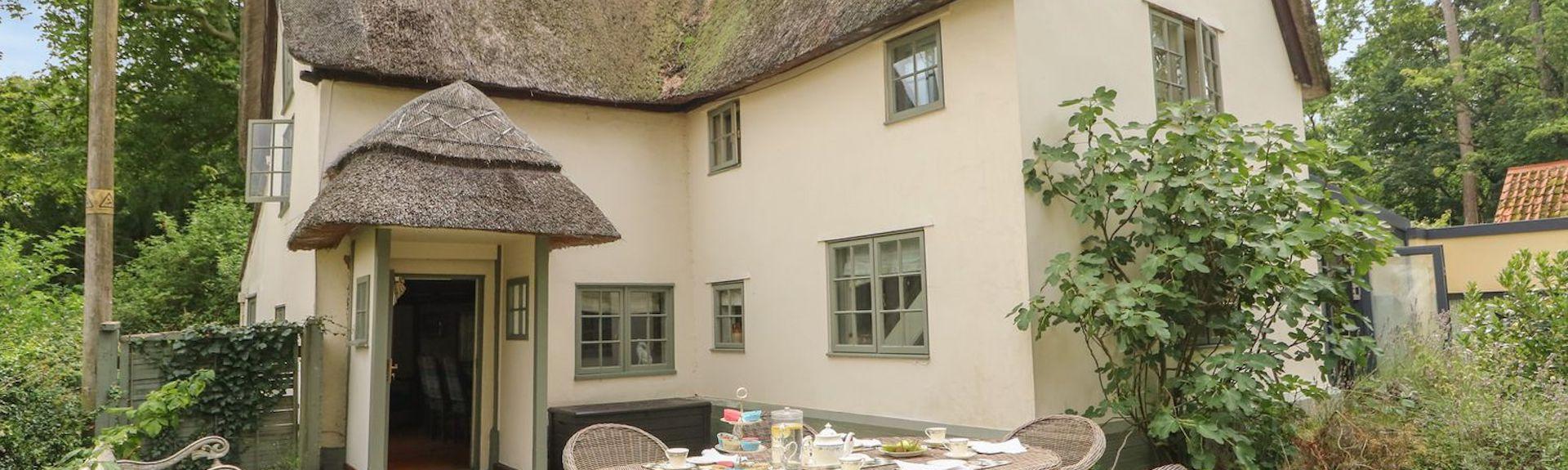 The extrior of  large thatched holiday cottage overlooking a patio garden with whicker picnic table and chairs
