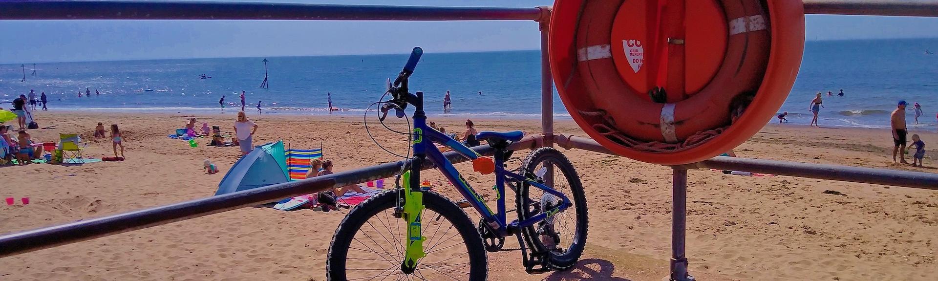 A bicycle is tethered to a railing alongside a life buoy overlooking a large sandy beach on which people are sunbathing. 
