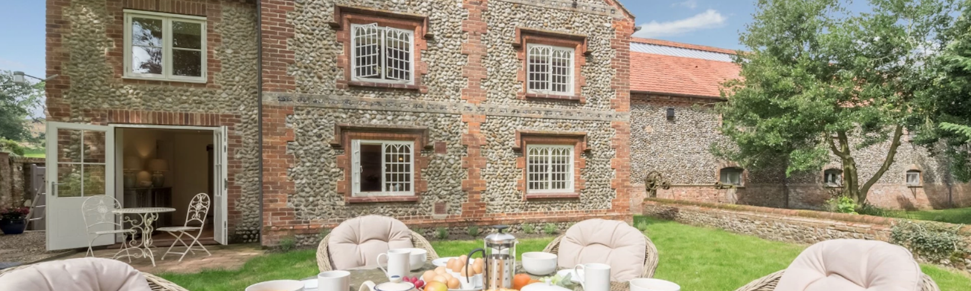 A large stone cottage buklt with pebble and brick oveerlooks a lawn on which is a wicker table and chairs laid for breakfast.