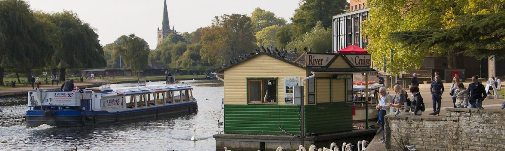A riveerside location with a river cruise boat. Ducks and swans are being fed from a stone, tree-lined treeside. 
