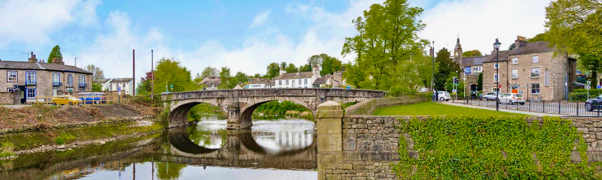 A twin-arched stone bridge is reflected in the still waters of the river below. Stone-bult houses line the riverside