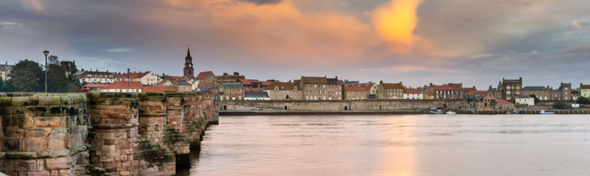 A waterside view at sunset in Berwick-upon-Tweed. The stone buttresses of anold bridge lead towards the far bank lined with old 3-storey houses.
