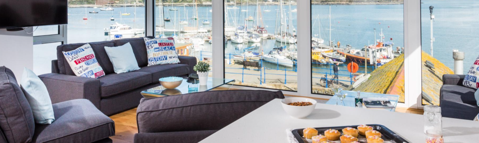 An open plan lounge with worktop and large comfortable sofas overlooks a harbourside view in Falmouth through floor-to-ceiling windows.