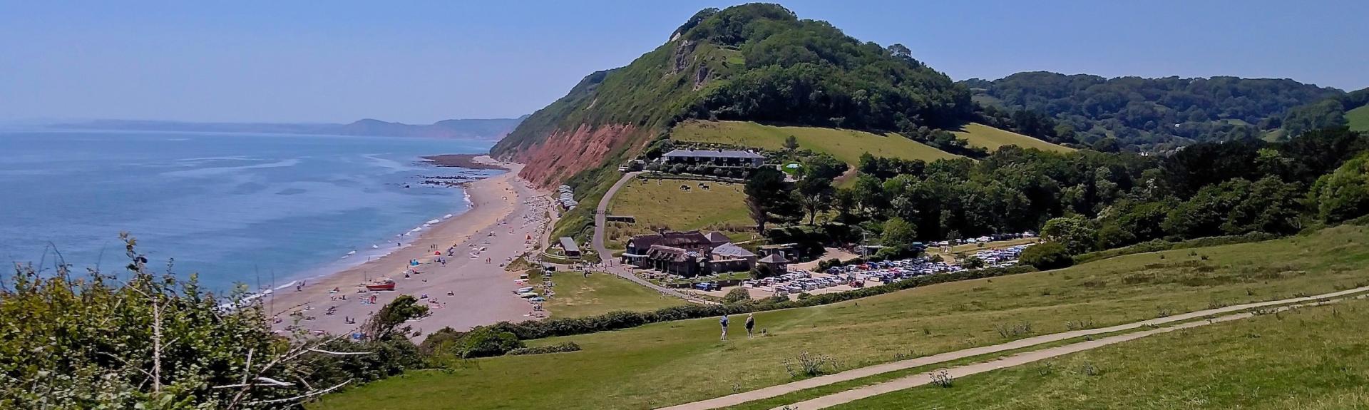 Branscombe beach in Devon. A field slopes down to a cluster of sea shanties overlooking a wide shingle beach, beyond which the lad rises to form cliffs topped by woodland. 