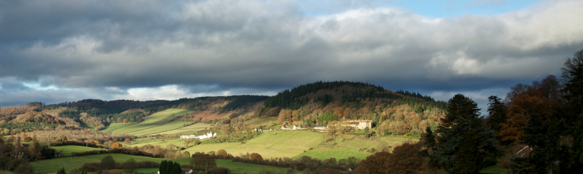 A landscape view of the Wye Valley with trees and open fields. In the foreground are some stone farm buildings  beyond which the land rises to a small hill on the horizon.