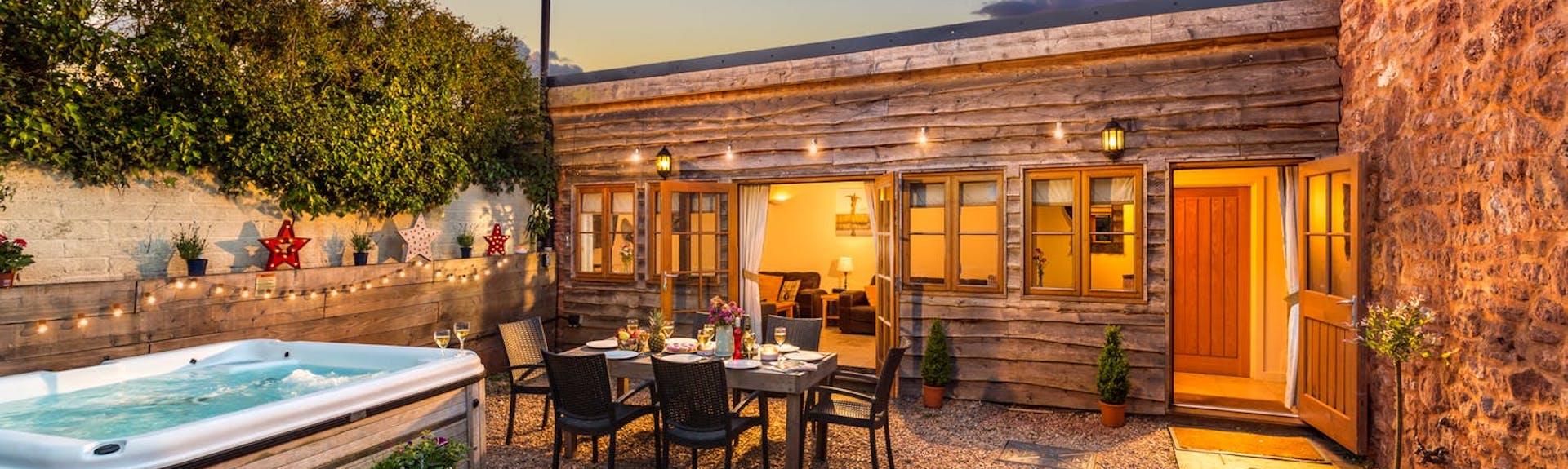 A wood-clad single storey holiday cottage at dusk overlooks a couryard with a table laid for dinner and a hot tub.
