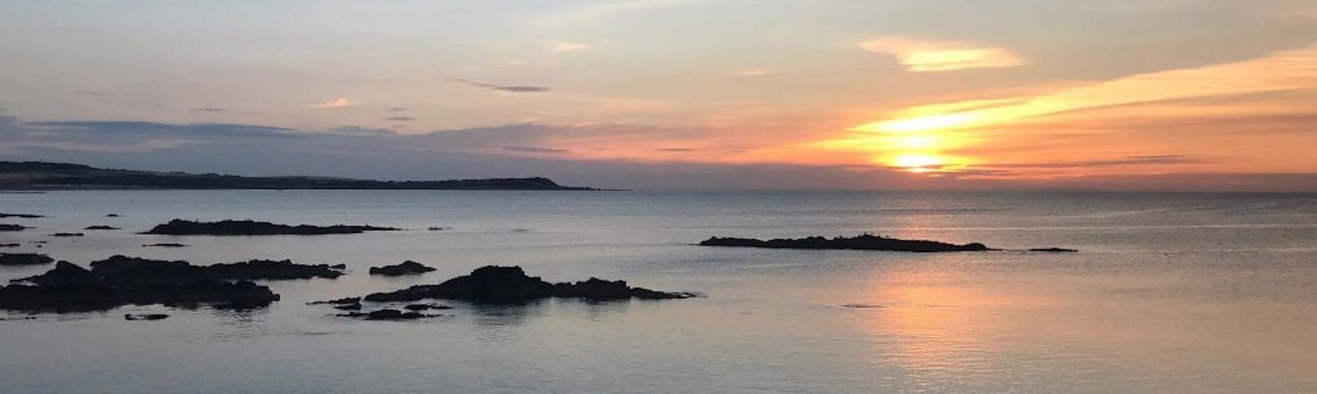 Sunset over The Moray Firth, a calm sea reflecting the sinking sun with tiny rocky islands silhouetted against the darkening sky.
