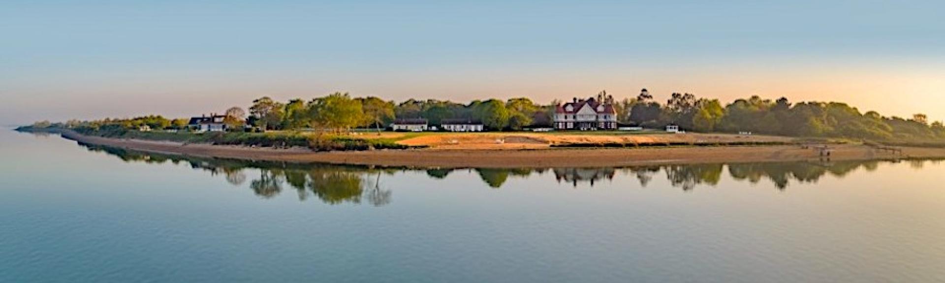 A low Island with trees. A handful of Essex holiday cottages and sandy beaches are reflected in a calm sea