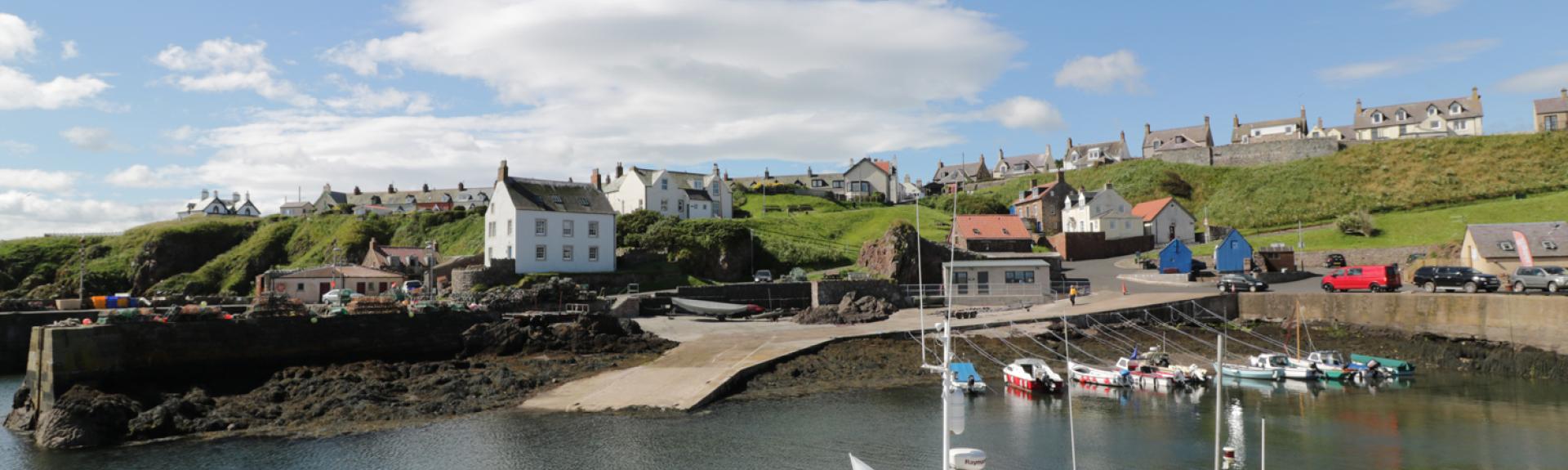 Small craft and fishing boats are moored to a quayside in St. Abbs, with village builldings in the backfround.