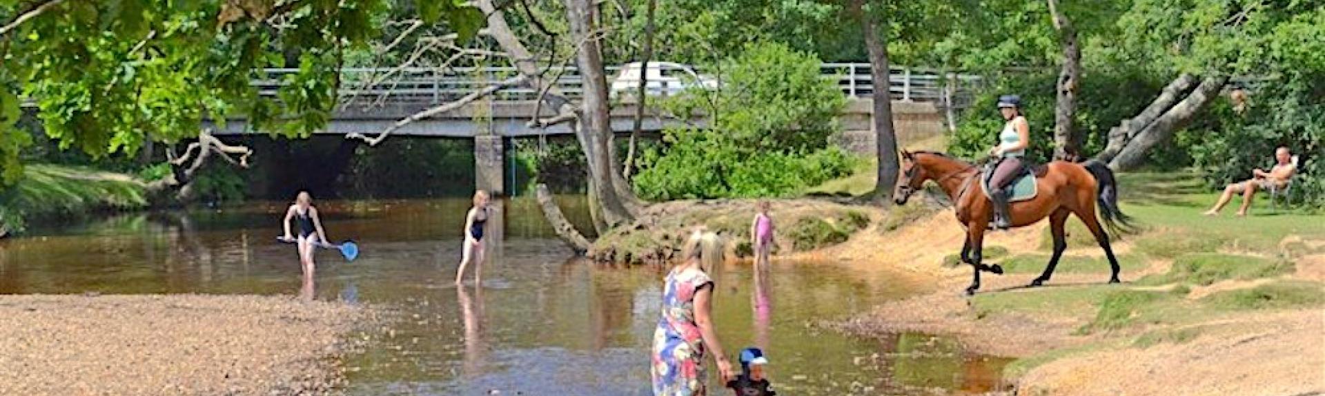 People paddle by a bridge in a New Forest stream as a rider on a horse crosses it.