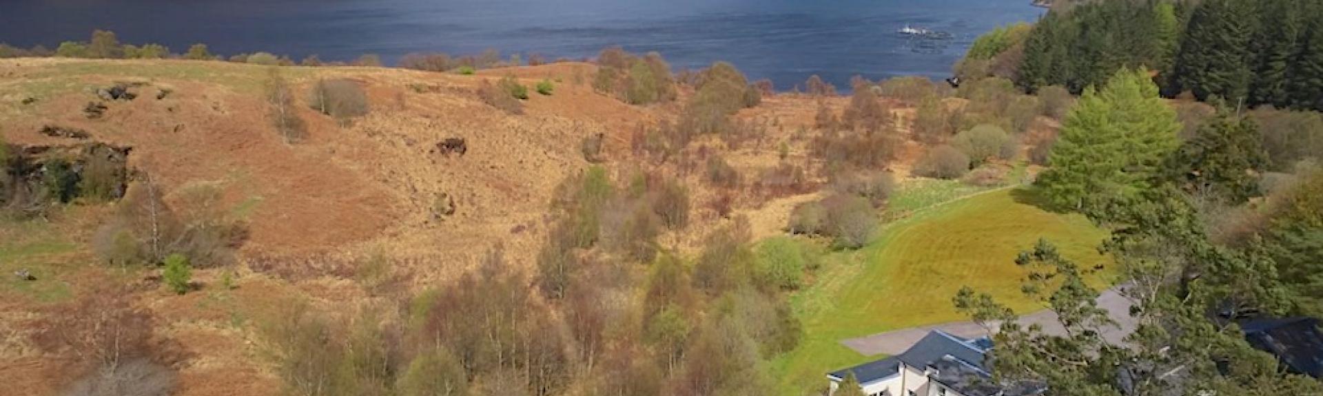 Aerial view of a cottage surrounded by open moorland with a loch in the background.