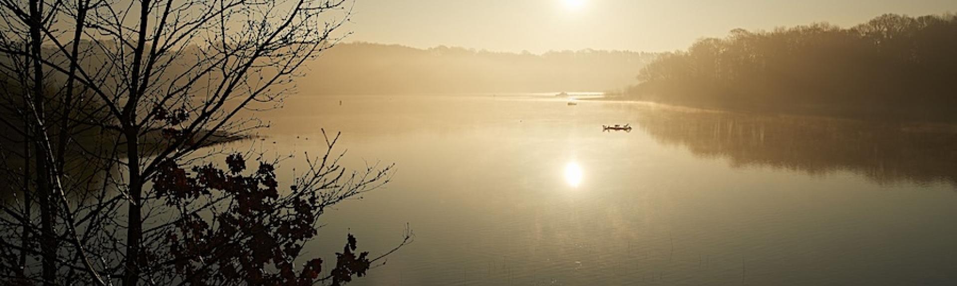 A tiny boat  is silhoutted in the setting sun on the flat calm surface of a large reservoir.
