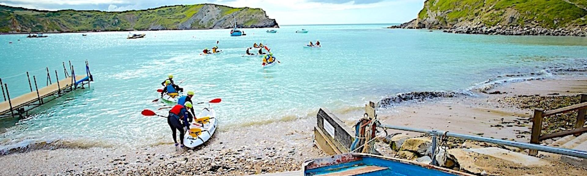 Kayaks paddle out to sea from a small Dorset beach on which a small wooden boat is anchored.