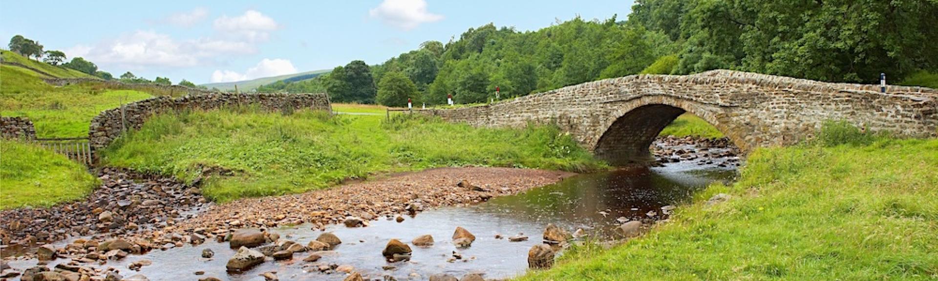 A single arched stone bridge crosses a shallow, rock-filled river in the countryside.