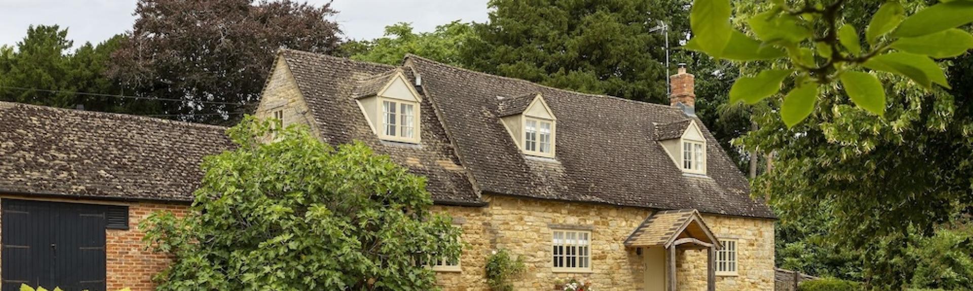 A Cotswold cottage with a back drop of tall tress sits above a large shingled drive