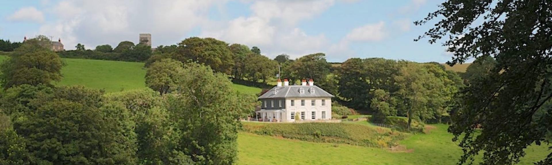 A Cornish country house glimpsed between trees overlooks a grassy hillside.