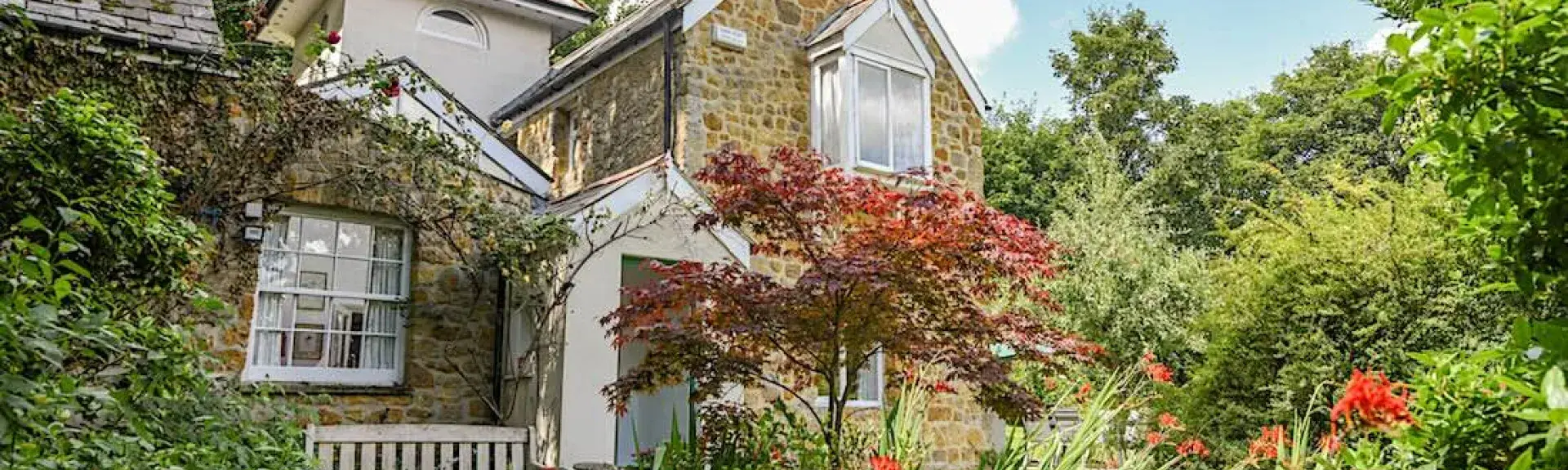 Exterior of a stone-built Dorset cottage overlooking a garden overflowing with shrubs and flowers.