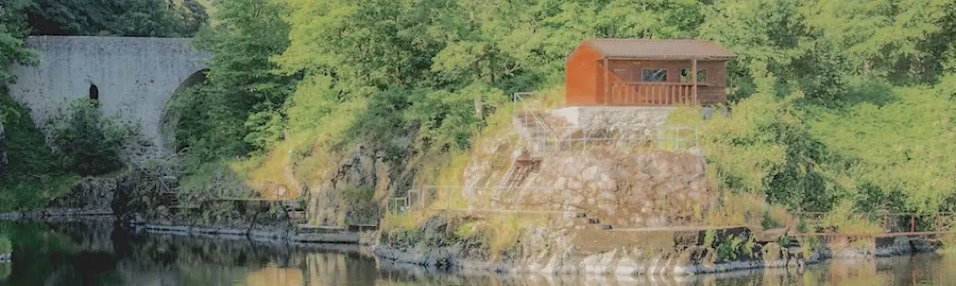 A wooden lodge in Aberdeenshire backed by trees is reflected in the calm waters of a river below it.