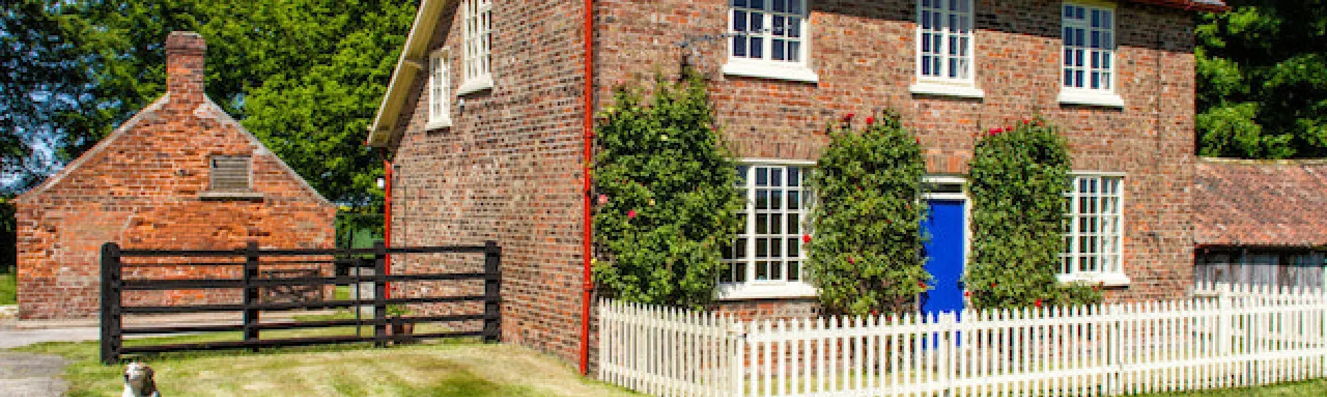 A double-fronted brick-built purpose-built detached cottage surrounded by a white picket fence lawns, and woodland.