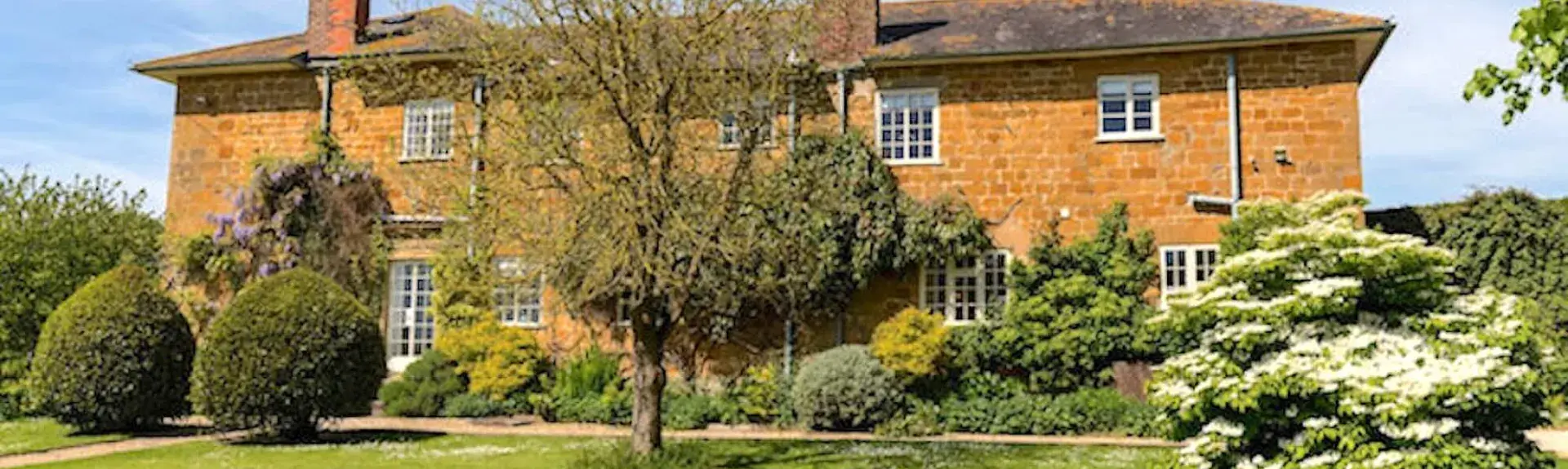 A 2-storey, Dorset golden-stone-built farmhouse overlooking a lawn surrounded by small trees, shrubs and flower borders.