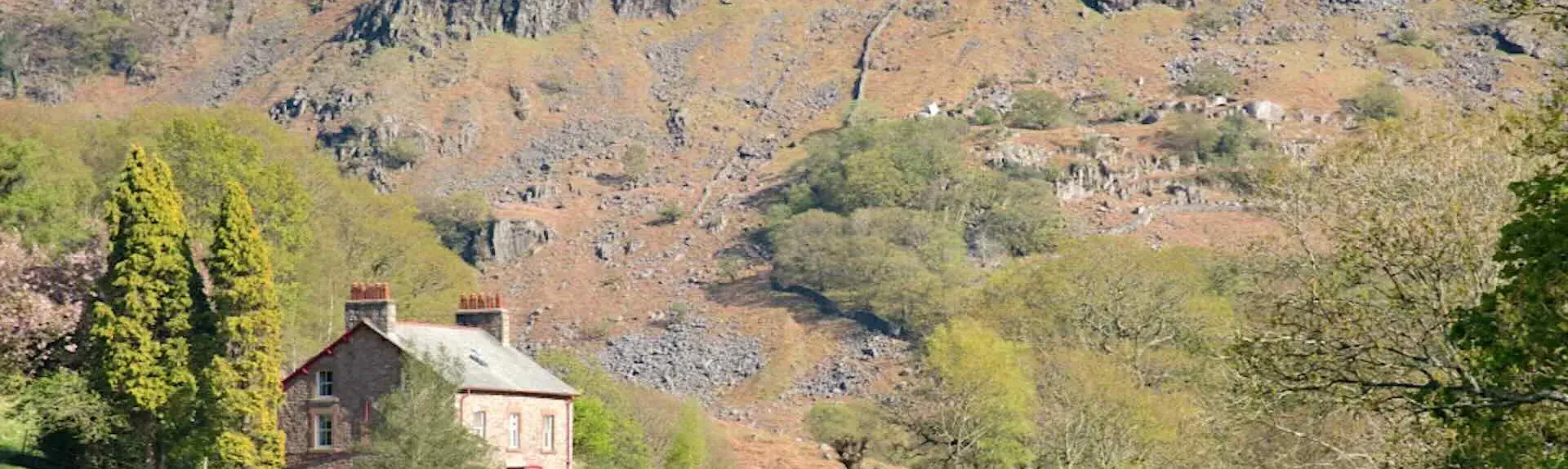 A solid-stone-built farmhouse nestles beneath a steep and rocky dcree-slope with stunted trees.