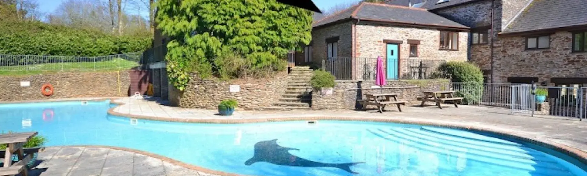 A wide terraace surrounds a kidney shaped open air pool overlooked by a large Devon holiday cottage and a lawn.