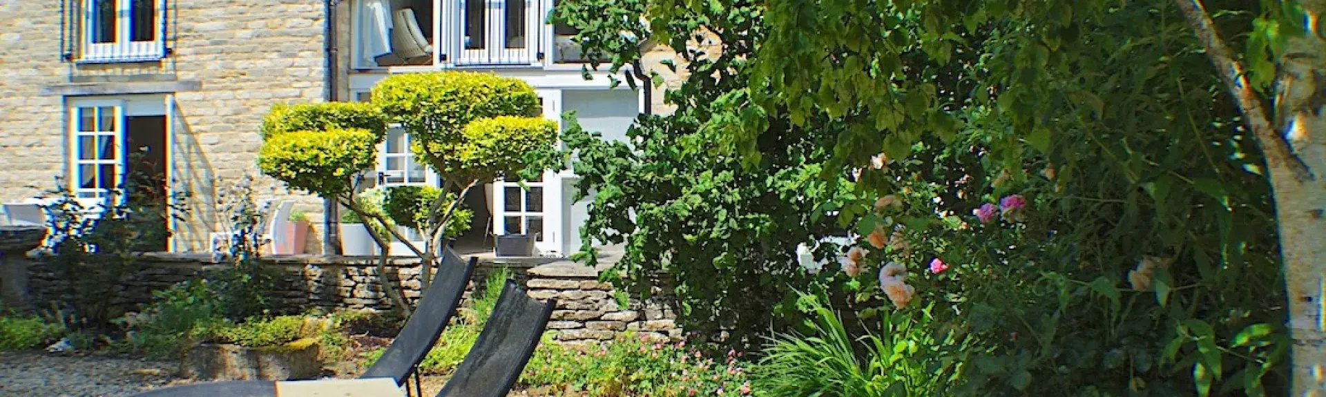 A Cotswold stone cottage with a long 1st floor balcony, half-screened by tall shrubs. 