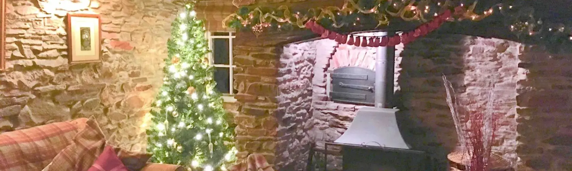 A stone-walled cottage lounge at Christmas with a Christmas tree and a large inglenook fireplace.