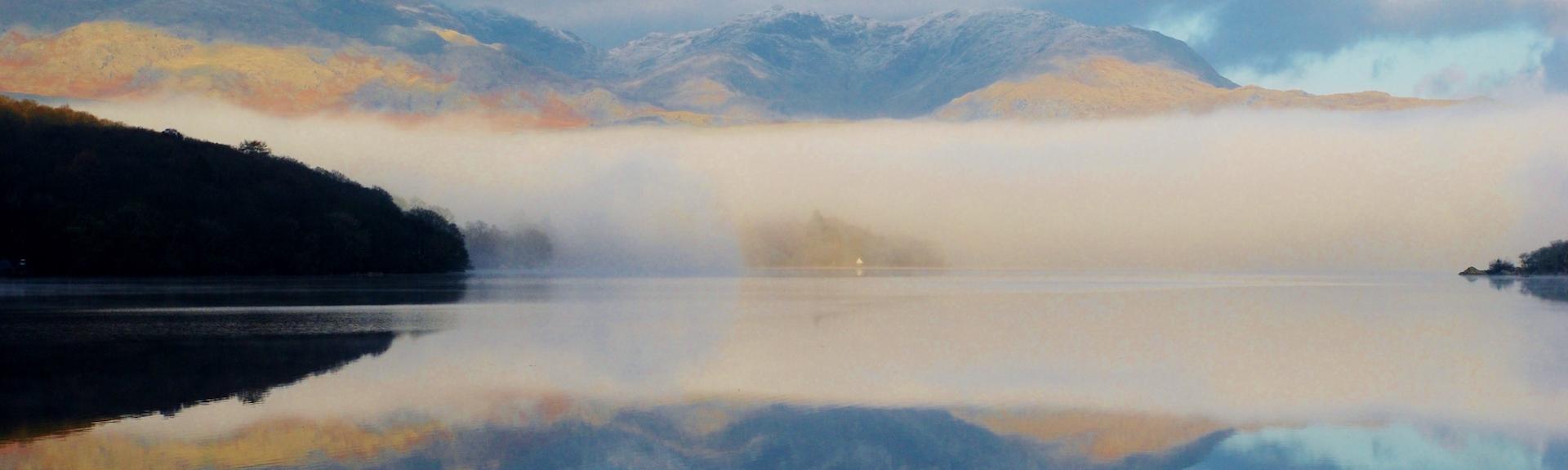 In the Lake District, a calm surface of a large lake reflects the fleecy cloud-filled sky above