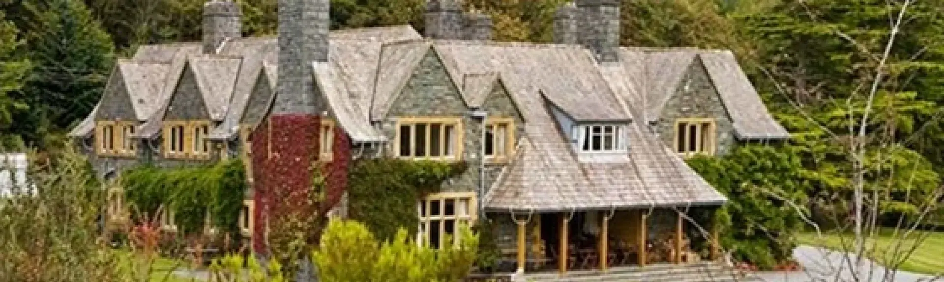 A large slate-roofed house house backed by tall trees overlooks a large lawn.