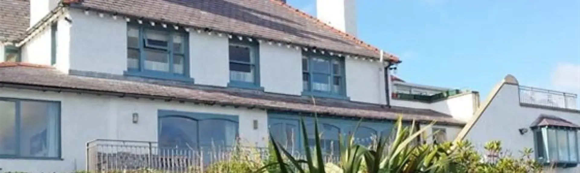 Exterior of a large holiday Anglesey holiday cottage with many windows  and terrace overlooking a shrub-filled garden.