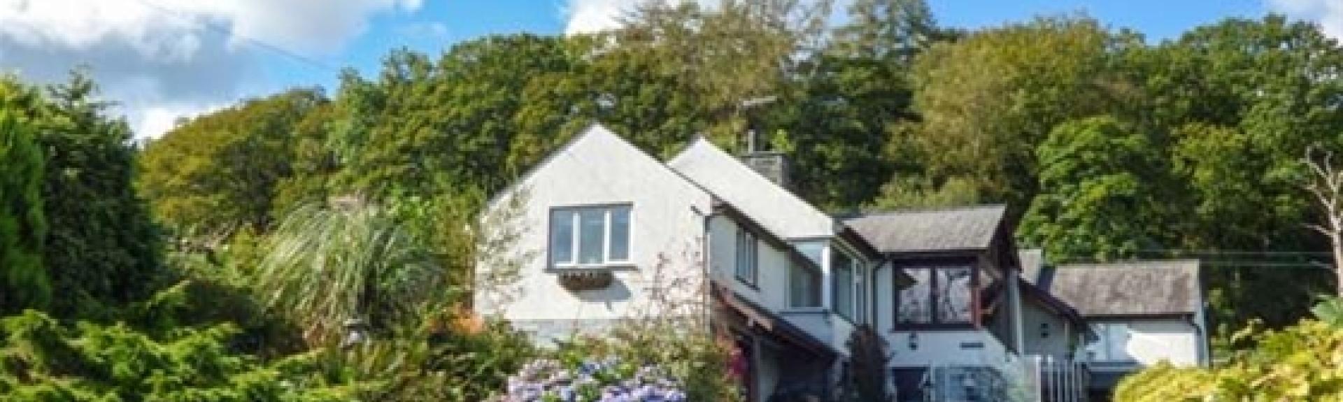 A large Coniston holiday home at the head of a hedge-lined tarmac drive with a mature woodland background.