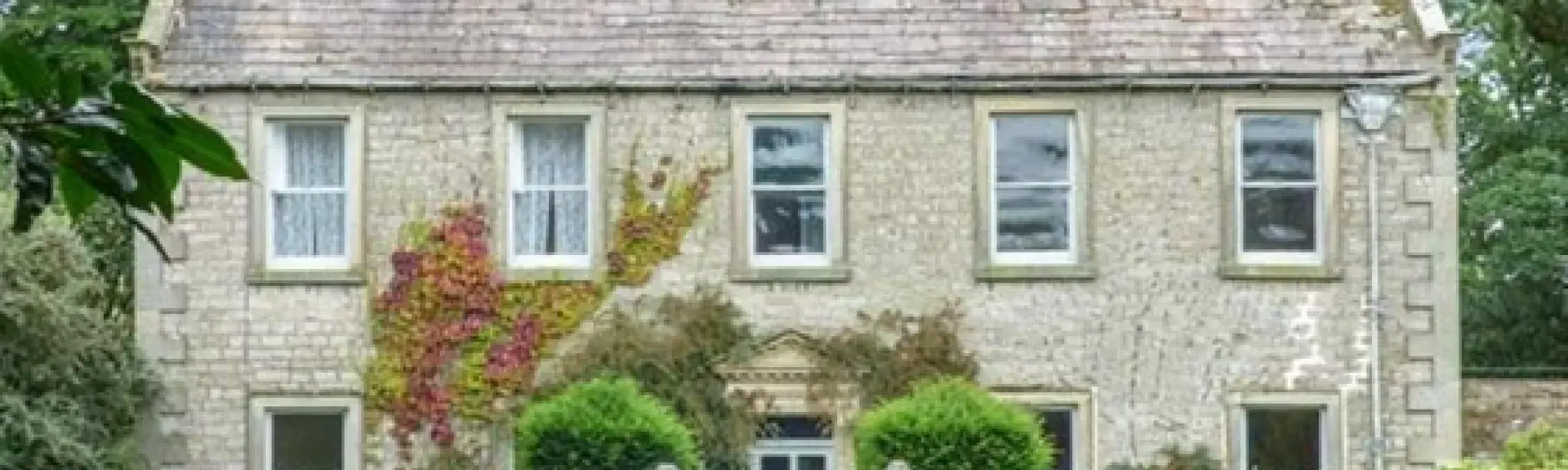 A classic country house with a row of five windows overlooks a shrub0filled garden lined by wrought iron railings. 