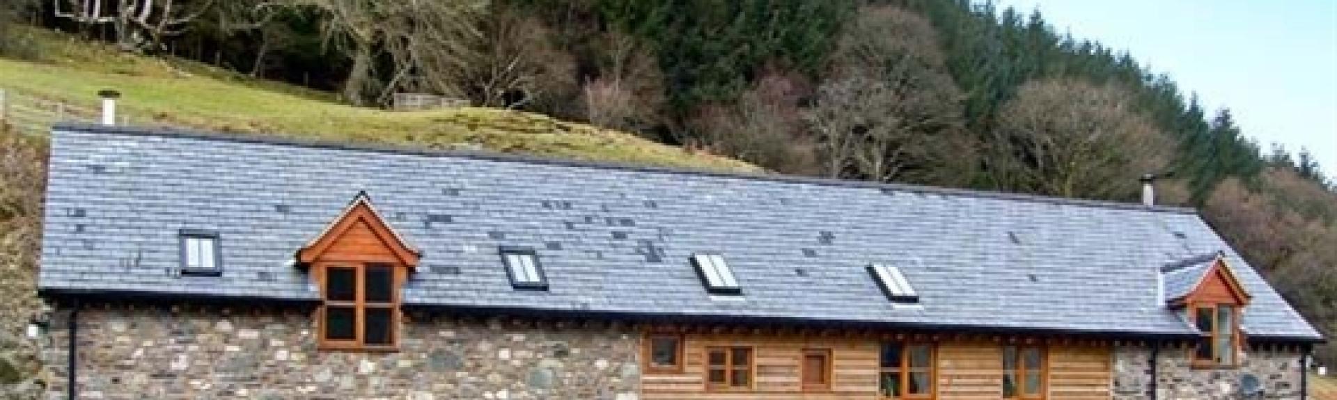 A slate-roofed, stone holiday cottage in Powys nestles beneath a step hill toppped with a wood.