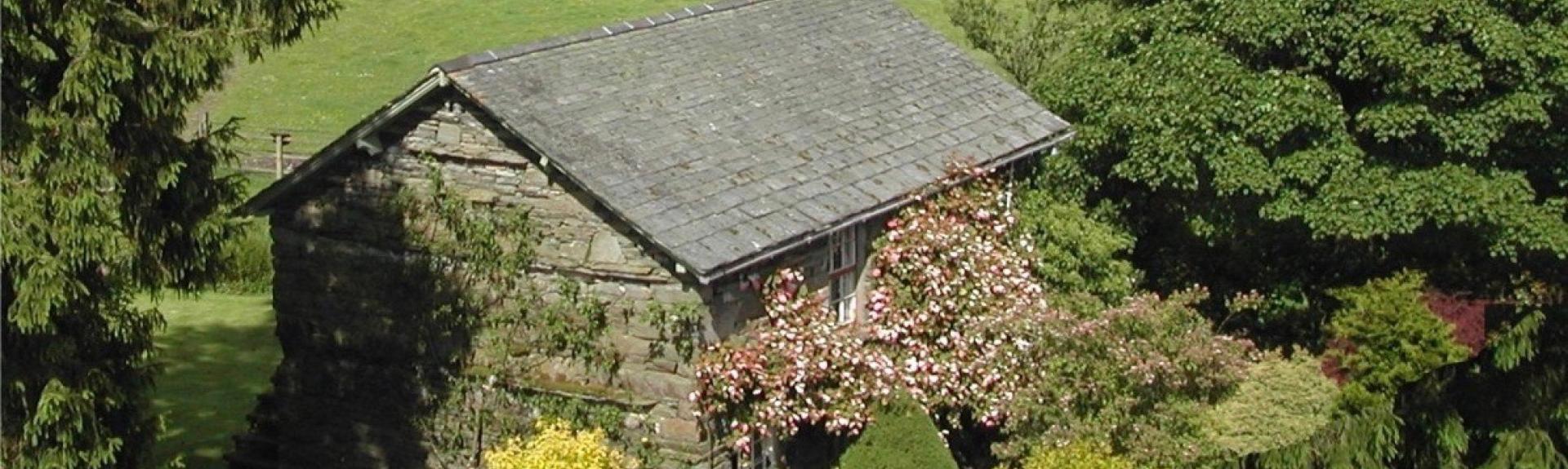A stone built, slate roofed Lake District holiday cottage surrounded by shrubs and small trees with hilly fields in the distance.
