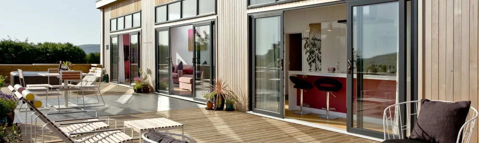 Exterior of a contemporary eco-lodge with large floor-to-ceiling plate glass windows and doors opening onto a wooden deck with a hot tub.