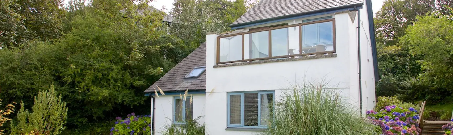 Exterior of a contemporary house in Cornwall with a balcony overlooking a large lawned garden bordered with shrubs and trees.