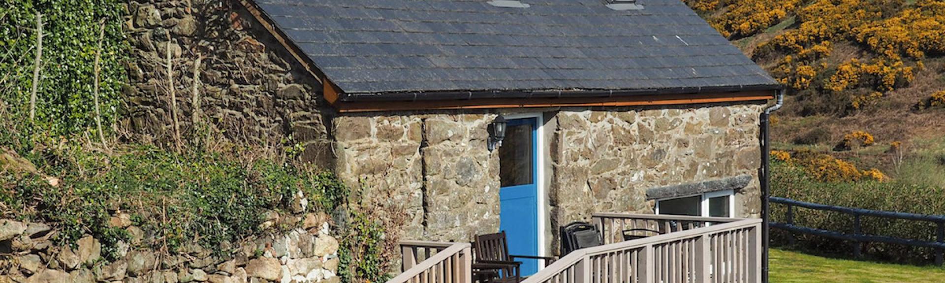 A barn conversion to create a holiday cottage in Abersoch: a wramp leasds to a wooden deck with outdoor seating and an entrance to this stone-built property with rural and sea views.