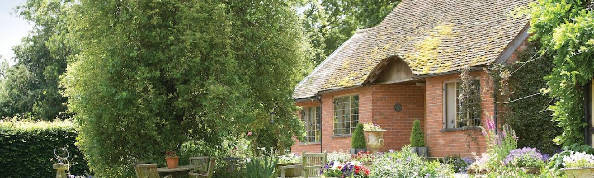 Exterior of s brick-built period garden cottage backed by mature woodland and in front of a lawn with flower-filled borders.