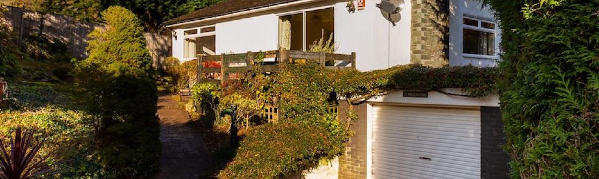 A modern house with a vine covered 1st floor balcony overlooks a shrub-filled front garden.