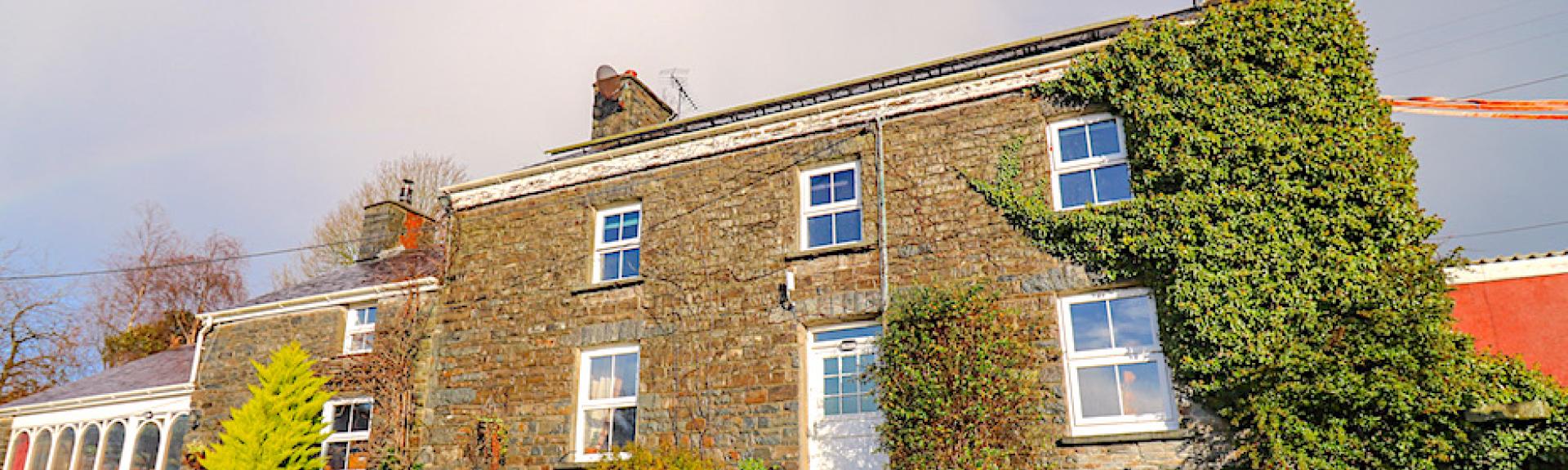 A double fronted, stone-built Welsh farmhouse sits on a flower-filled raised bank above a lane.