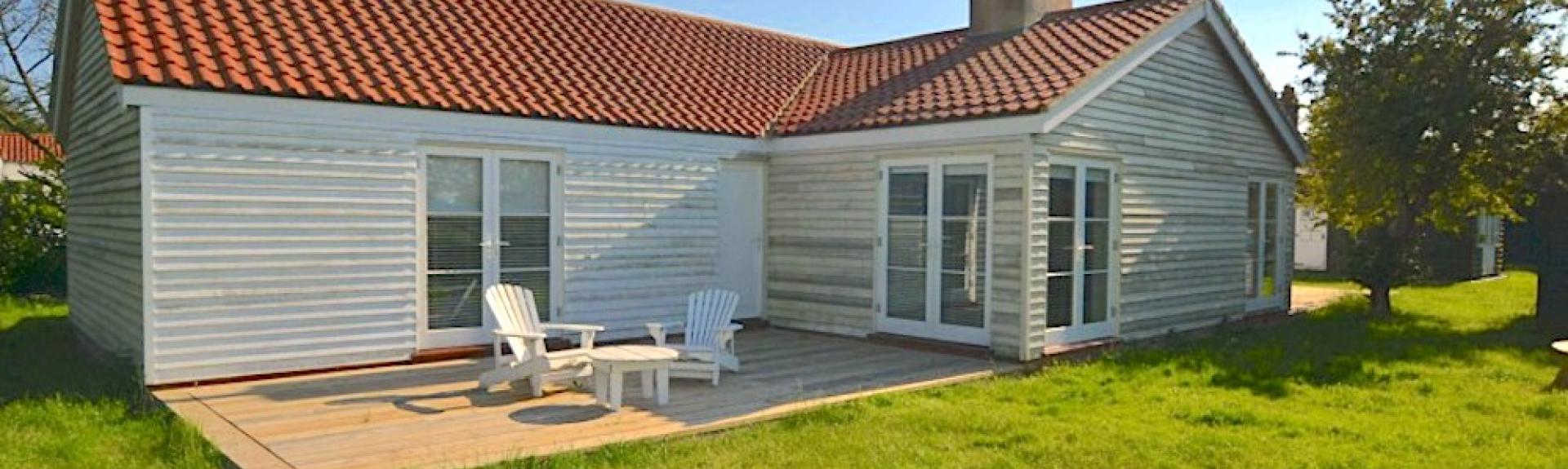 An L-shaped, wood-panelled Essex holiday bungalow with a wooden deck containing a table and sunlounger all overlooking a large lawn.