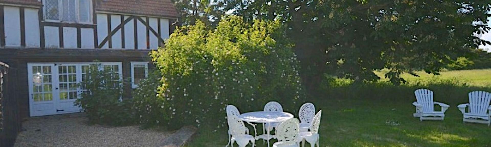 Outdoor dining chairs cluster around a table on a tree-lined lawn on Osea Island. Behind them is a cross-timbered holiday cottage on Osea Island.