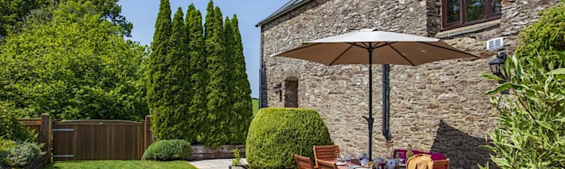 A table with parasol and chairs tands on a sunny patio behind a stone built holiday cottage in Devon.
