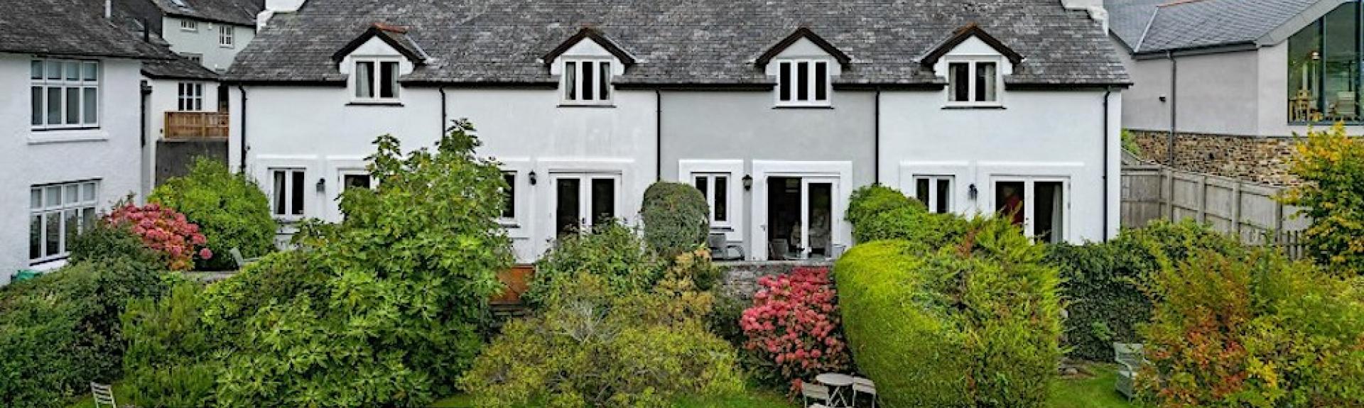 A terrace of 4 slate-roofed cottages over looking a lawn with tall shrubs and an open field in Dittisham. 