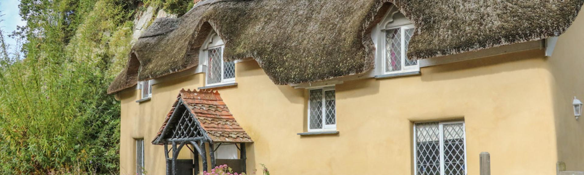 Exterior of a thatched cottage with porch and low stone wall.